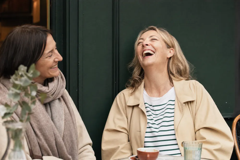 Two women in their forties sitting outside a cafe, one laughing freely